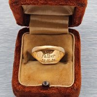 A yellow gold signet ring engraved with 'From Father 26-10-24' resting inside an open orange-brown velvet jewellery box on a light gray textured background.