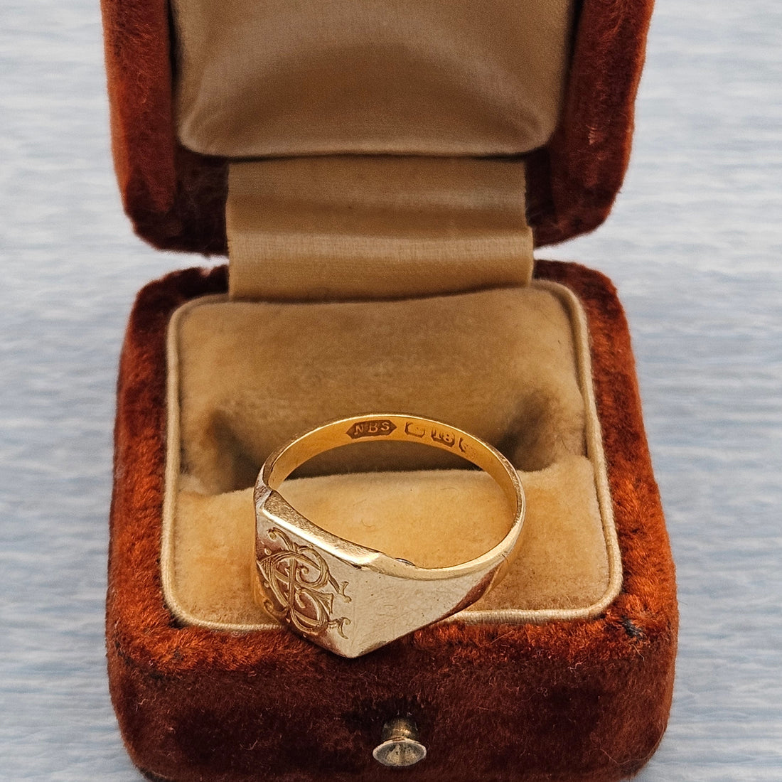 A gold signet ring with an engraved pattern and hallmarks sits inside an open brown velvet ring box with a tan satin lining, set against a light gray textured background.