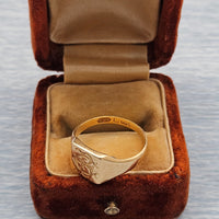 A gold signet ring with an engraved pattern and hallmarks sits inside an open brown velvet ring box with a tan satin lining, set against a light gray textured background.