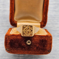 A vintage gold signet ring with an ornate monogram design, displayed inside an open antique brown velvet ring box on a light gray textured background.