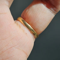 A close-up of the back of thin gold band ring worn on a finger, showing the textured metal against skin and a dark gray background.