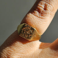 A close-up of a gold signet ring with an engraved monogram and patina (including many surface scratches and nicks), worn on a finger.