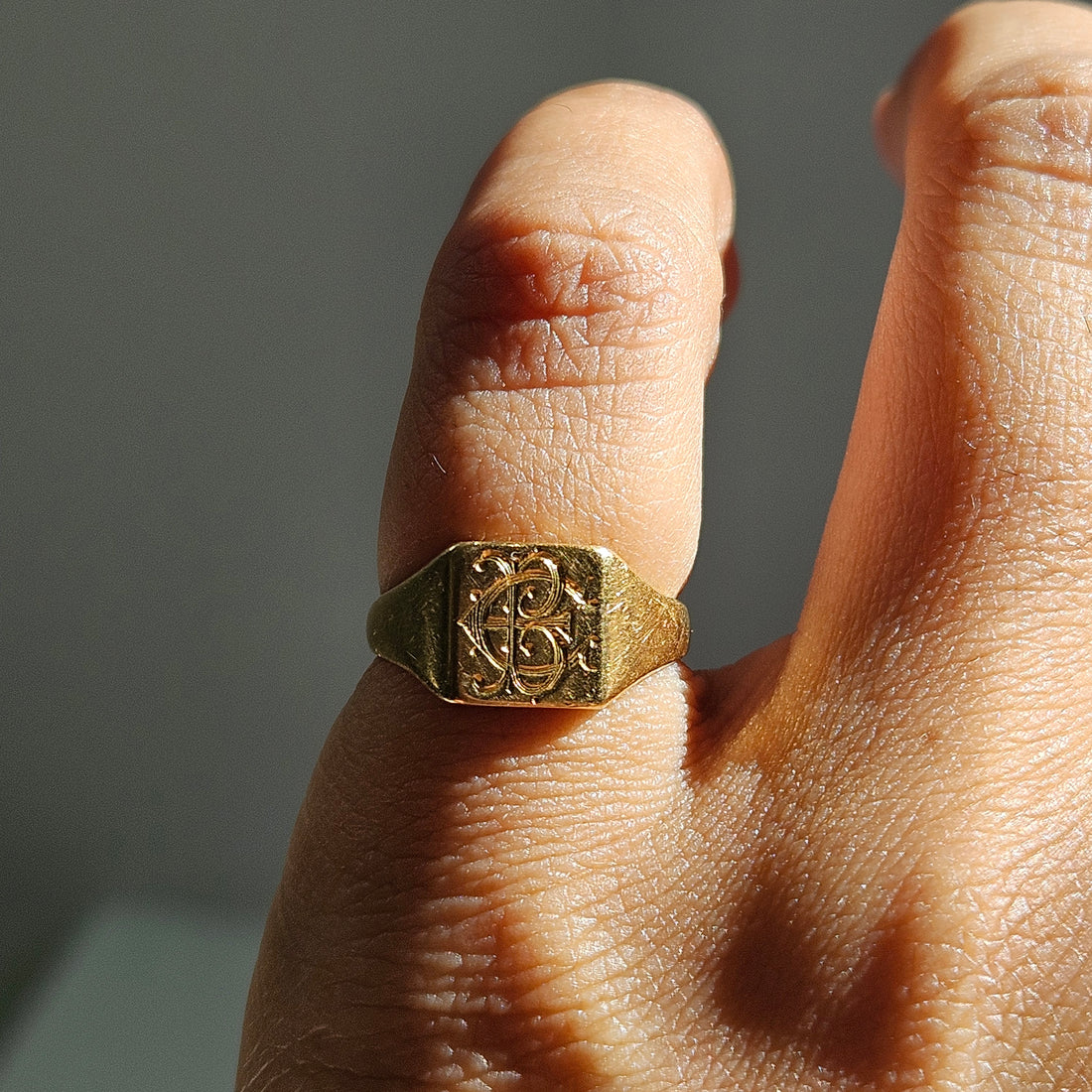 A close-up of a hand wearing a gold signet ring with an engraved monogram design on a square face, set against a neutral gray background.