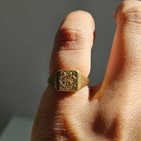 A close-up of a hand wearing a gold signet ring with an engraved monogram design on a square face, set against a neutral gray background.